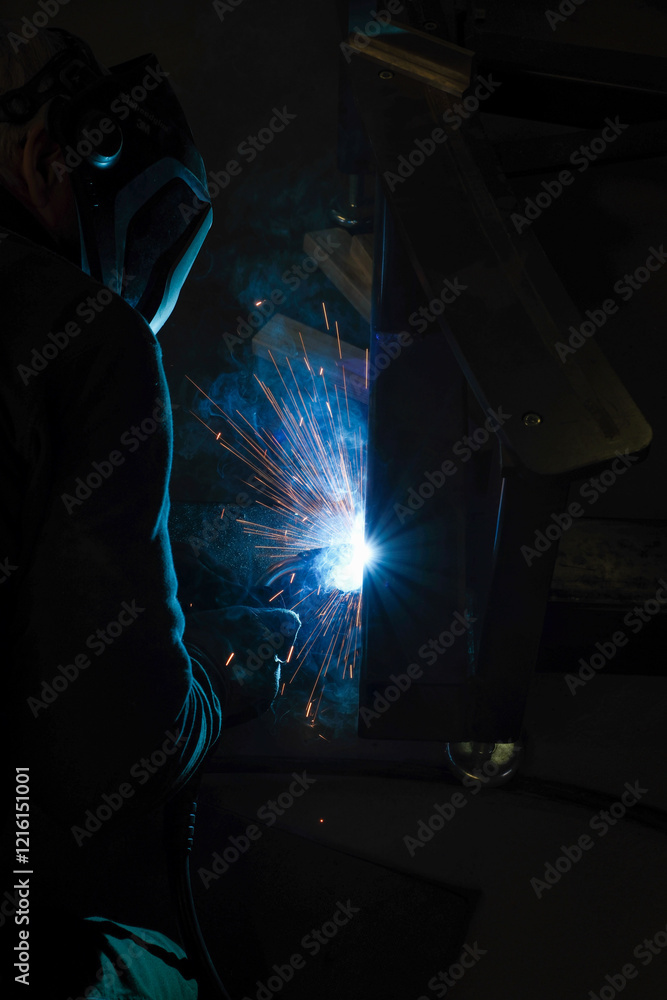 a person wearing a welding mask works on metal with a welding torch, creating sparks through the welding process