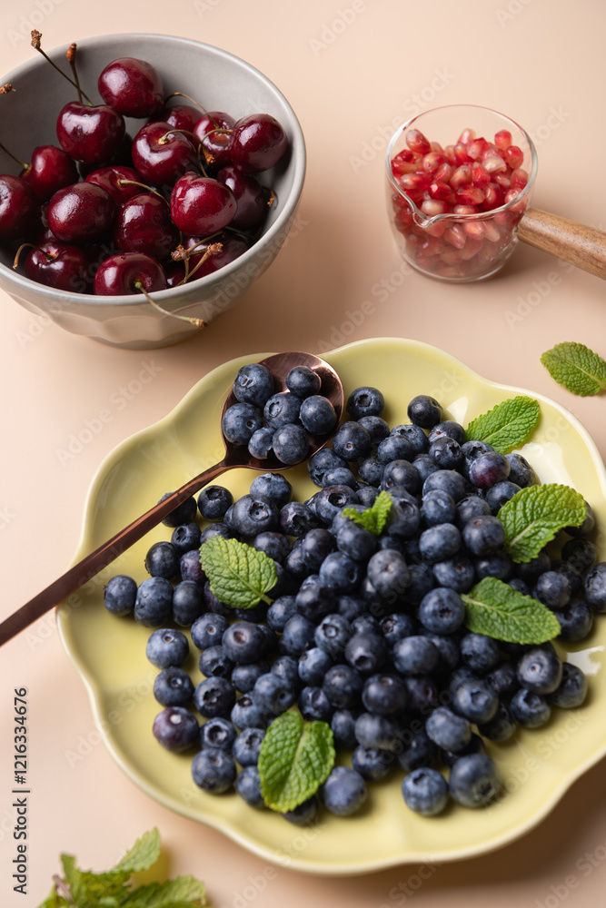 Blueberries, cherries and pomegranates on plate and containers, on light background and decorated with mint leaves. Natural cooking and healthy eating concept Blueberries, cherries and pomegranates on plate and containers, on light background and decorated with mint leaves. Natural cooking and healthy eating concept
