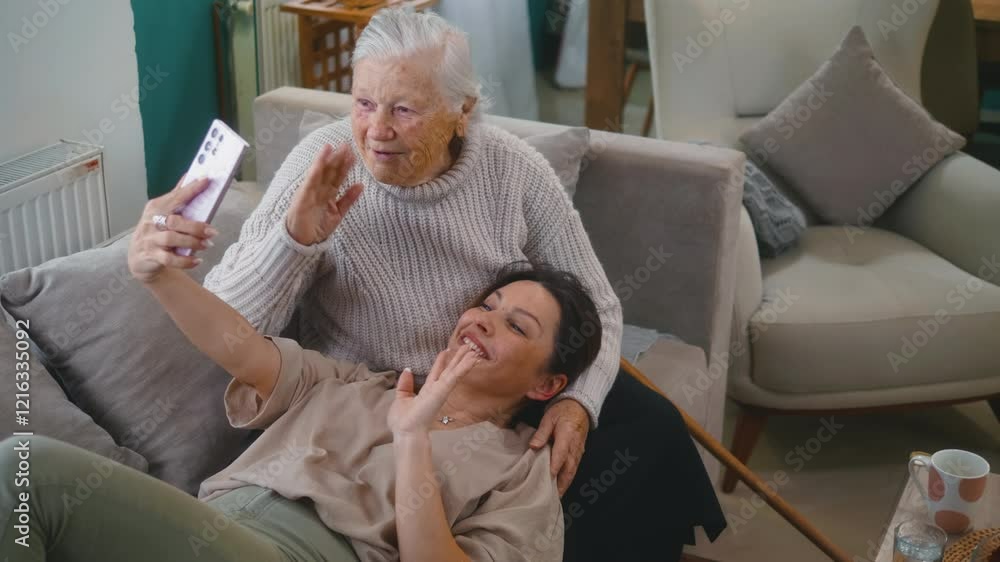 Adult daughter and her senior 80s mother laughing while having fun on the phone at home, having a video call, using funny apps, and taking selfies for social media