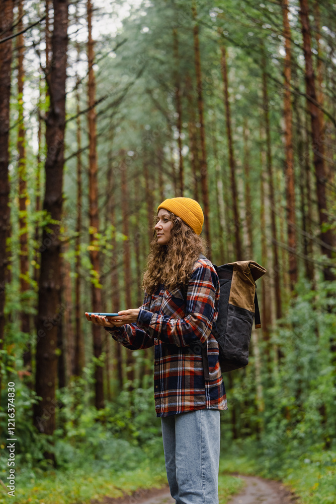Fototapeta premium Young female hiker with backpack and yellow beanie using smartphone gps app for navigation on forest trail, exploring nature and enjoying outdoor adventure
