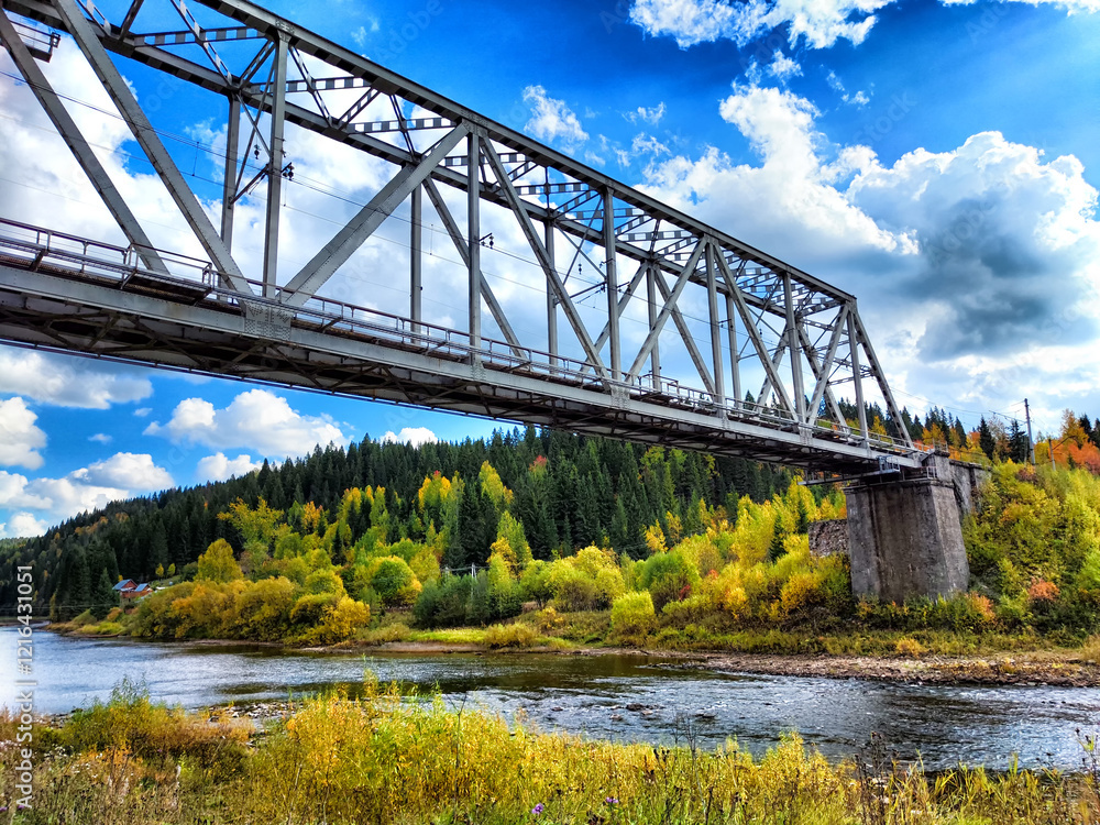 Obraz premium Metal bridge arches over a river surrounded by colorful autumn foliage under a bright blue sky