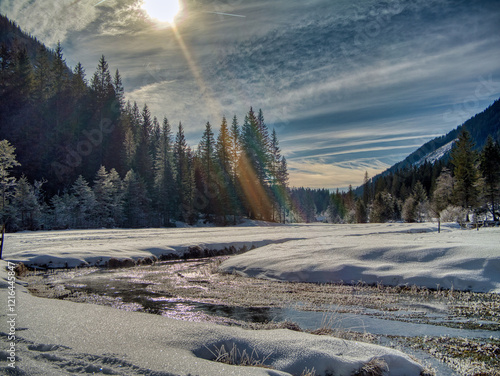 Winterliche Flusslandschaft im Sonnenschein
Ein glitzernder Fluss schlängelt sich durch eine verschneite Winterlandschaft, umgeben von Wäldern und Bergen, während die Sonne durch die Bäume strahlt. photo