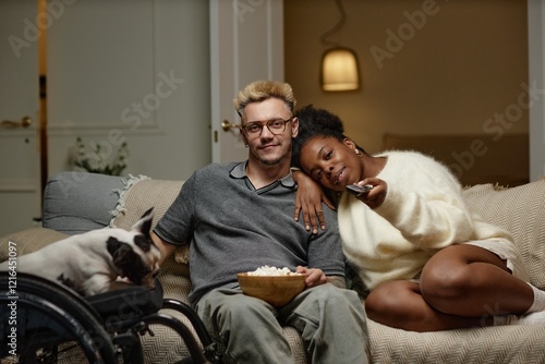 Portrait of calm male wheelchair user sitting on couch with African American girlfriend holding remote control while watching classic films on romantic home date, hygge concept, copy space photo