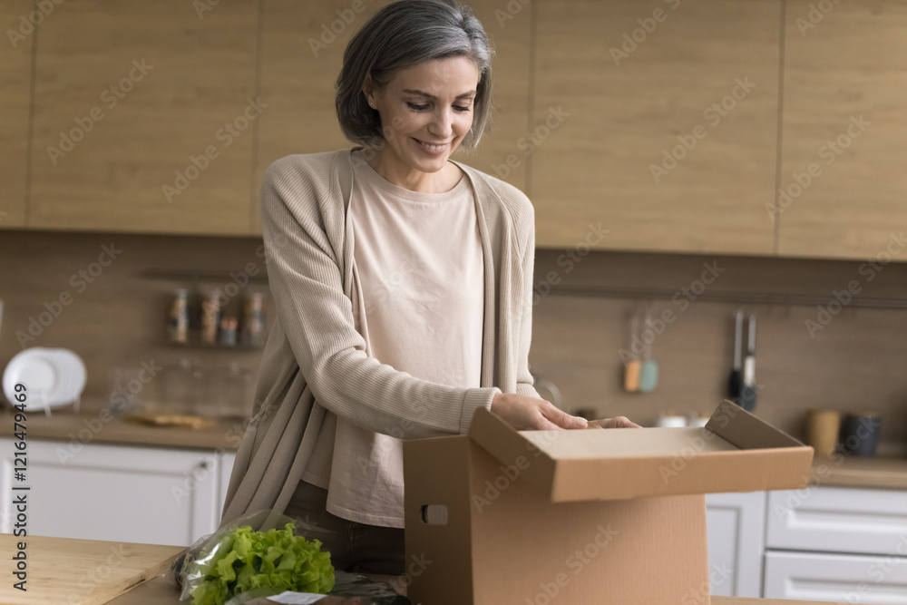 Home food delivery. Smiling middle aged woman housewife satisfied grocery store customer unpacking postal parcel box with fresh natural organic healthy food products ordered online at domestic kitchen Home food delivery. Smiling middle aged woman housewife satisfied grocery store customer unpacking postal parcel box with fresh natural organic healthy food products ordered online at domestic kitchen