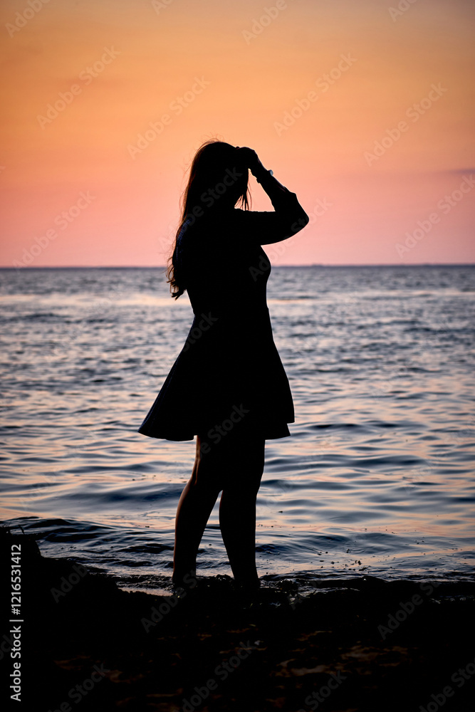 Fototapeta premium Vertical photo of a silhouette of a girl on the seashore against the background of the sunset and water