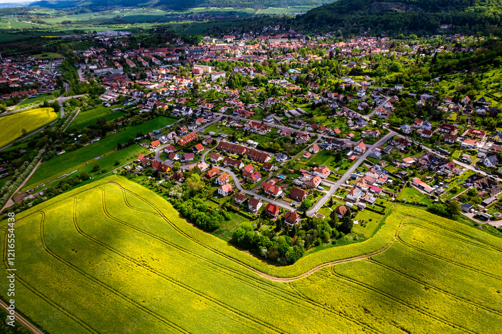 Top view of a field of yellow rapeseed flowers with a small town in the background. The sky is cloudy and the sun is shining through the clouds. Germany.
