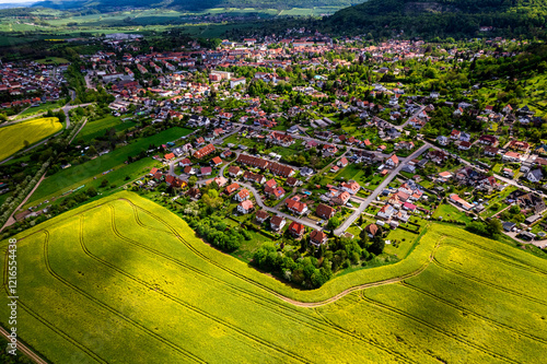 Obraz na płótnie Top view of a field of yellow rapeseed flowers with a small town in the background. The sky is cloudy and the sun is shining through the clouds. Germany. w salonie
