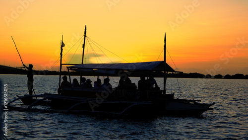 A silhouette of a traditional Philippine boat carrying passengers, gliding over tranquil waters beneath a vibrant sunset, evoking a serene and scenic atmosphere.