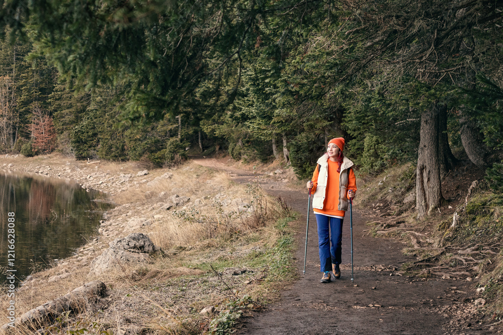 Obraz premium Hiker walking using trekking poles along forest trail near lake in autumn
