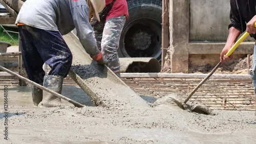 A team of construction workers is collaborating to shovel cement mix from a transport truck to build a concrete road under the hot sun