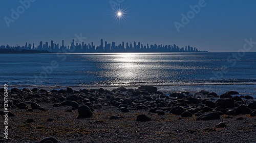Sun setting over city skyline reflected in calm ocean water with rocky shore in foreground.