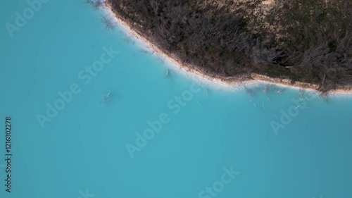 A turquoise tailings pond near a cement factory, surrounded by rugged hills. The vivid water contrasts with the barren land, creating a striking yet industrial scene of environmental impact photo