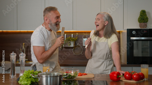 Funny Caucasian couple in love mature middle-aged senior old wife and husband retired married family singing song at spoons kitchenware joyful laughing woman man happy cooking healthy food in kitchen photo