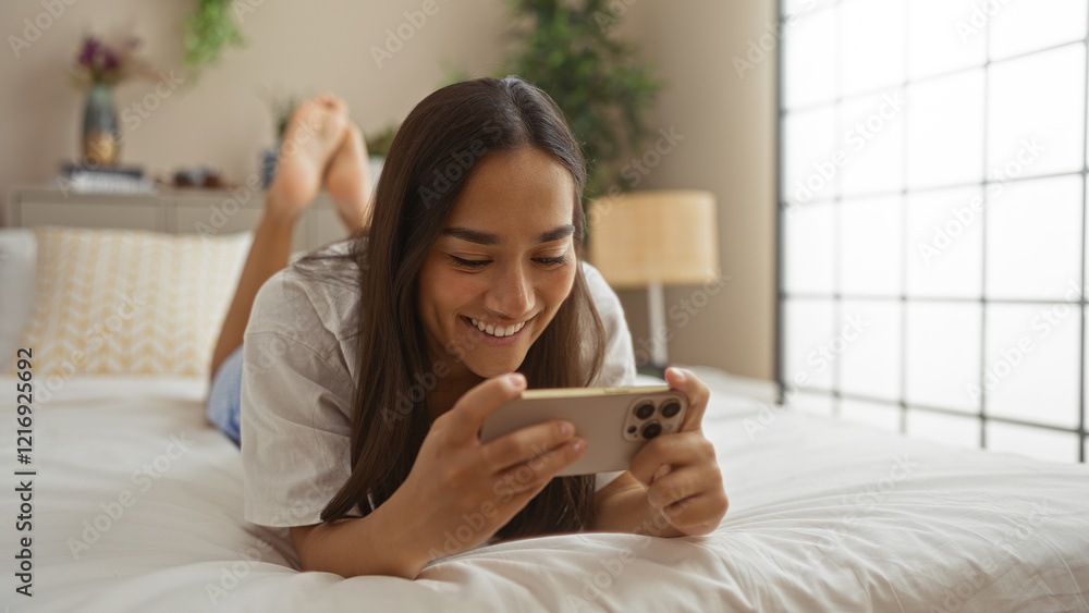 Young woman lying on bed in bedroom using smartphone, smiling happily in cozy home interior Young woman lying on bed in bedroom using smartphone, smiling happily in cozy home interior