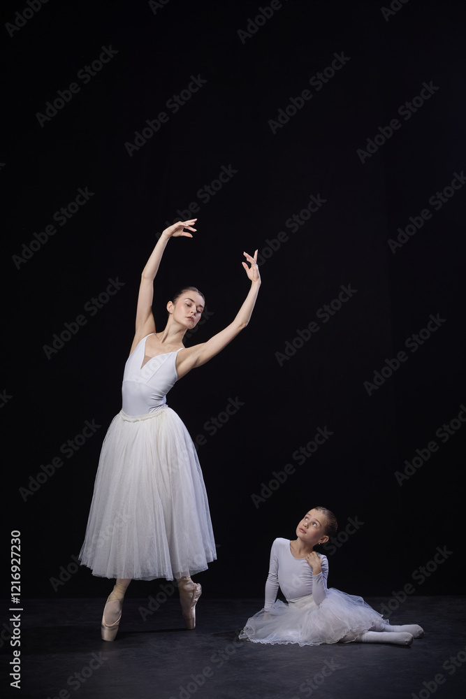 Adult ballerina posing with arms elegantly curved above head, child sitting nearby gazing upwards, both in white costumes, against black background. Concept of ballet, elegance and beauty, grace. Ad Adult ballerina posing with arms elegantly curved above head, child sitting nearby gazing upwards, both in white costumes, against black background. Concept of ballet, elegance and beauty, grace. Ad