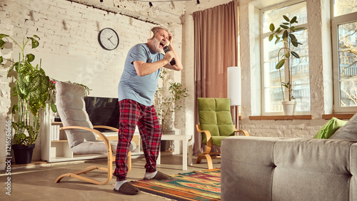 Elderly man in pajamas listening to music in headphones and emotionally singing in tv controller in warmly lit living room with plants, chair and large tv. Concept of happiness, elderly lifestyle photo