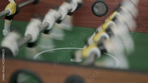 After goal in kicker teenager takes ball out of special hole and puts it into game, static shot. Friends play table football and actively rotate handles with figures of football players.