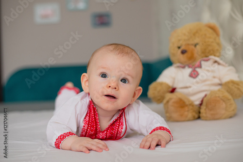 beautiful child boy lying on bed in Ukrainian embroidered shirt with teddy bear toy