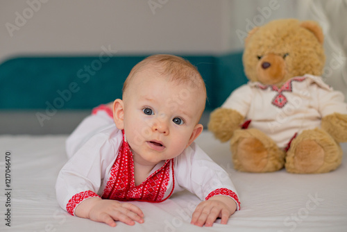 beautiful child boy lying on bed in Ukrainian embroidered shirt with teddy bear toy