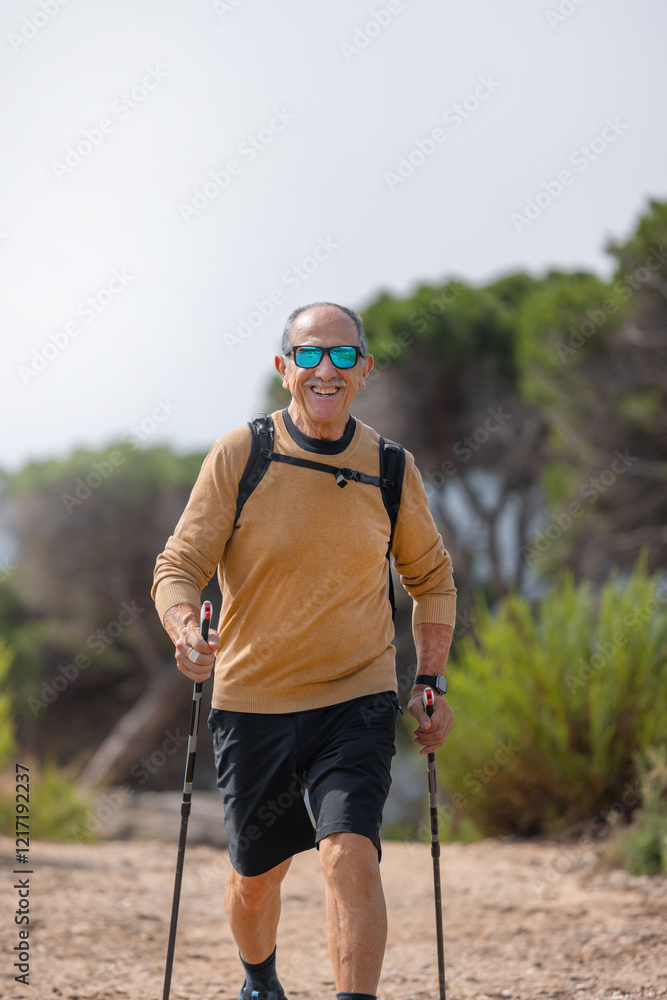 Smiling senior man looking at the camera with sunglasses and sportswear, trekking with poles on a dirt path. Surrounded by green bushes, pine trees, and a bright sunny sky. Waist-up. Vertical Smiling senior man looking at the camera with sunglasses and sportswear, trekking with poles on a dirt path. Surrounded by green bushes, pine trees, and a bright sunny sky. Waist-up. Vertical