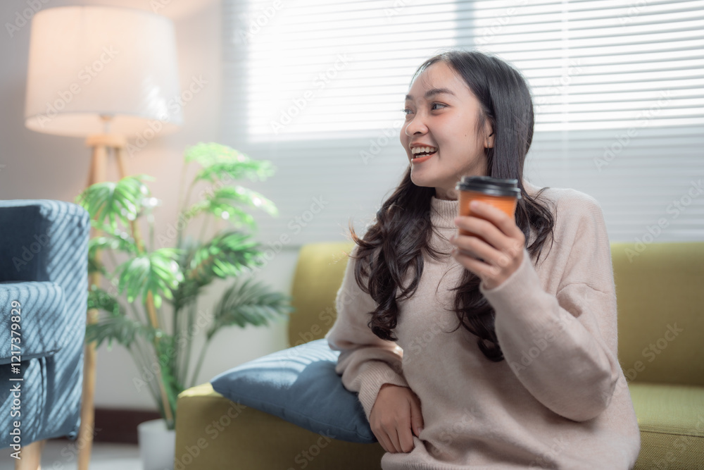 Happy young Asian woman relaxing on a cozy sofa, savoring a takeaway paper cup of coffee while enjoying her leisure time in a peaceful living room at home Happy young Asian woman relaxing on a cozy sofa, savoring a takeaway paper cup of coffee while enjoying her leisure time in a peaceful living room at home