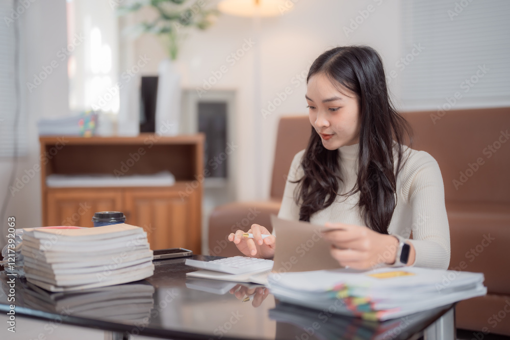 Young focused asian businesswoman sitting at home office desk and making calculations using calculator while reviewing domestic expenses and invoices Young focused asian businesswoman sitting at home office desk and making calculations using calculator while reviewing domestic expenses and invoices