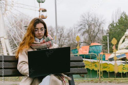 A young woman with glasses and bright hair, researching in an abandoned city amusement park with a laptop. Ideal for themes of urban exploration, remote work, and unique work environments. photo