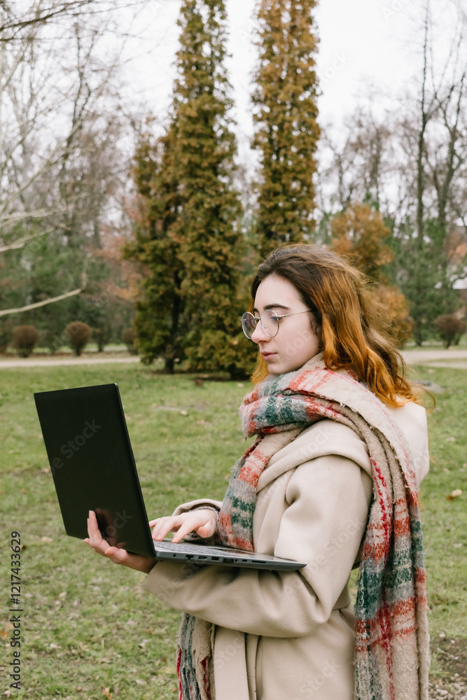 A young woman with glasses working or studying on her laptop in a city park on a cool, cloudy day. Perfect for themes of remote work, learning, and productivity in an urban setting. A young woman with glasses working or studying on her laptop in a city park on a cool, cloudy day. Perfect for themes of remote work, learning, and productivity in an urban setting.