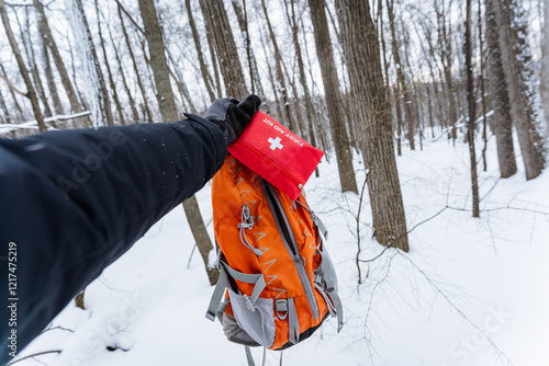 A person holds a bright red first aid kit above an orange backpack in soft snow, set against a stunning winter landscape filled with icy flakes and invigorating cold air photo