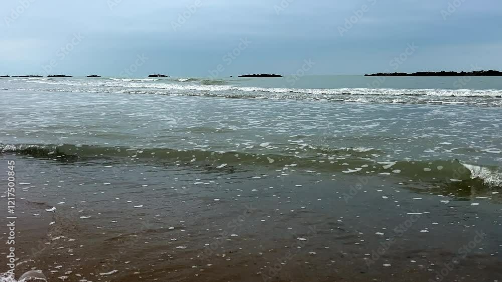 A scenic view of gentle foamy waves rolling onto the sandy shores of Pescara, Italy. The cloudy sky and calm waters create a peaceful coastal atmosphere, with rocky breakwaters visible in the distance A scenic view of gentle foamy waves rolling onto the sandy shores of Pescara, Italy. The cloudy sky and calm waters create a peaceful coastal atmosphere, with rocky breakwaters visible in the distance