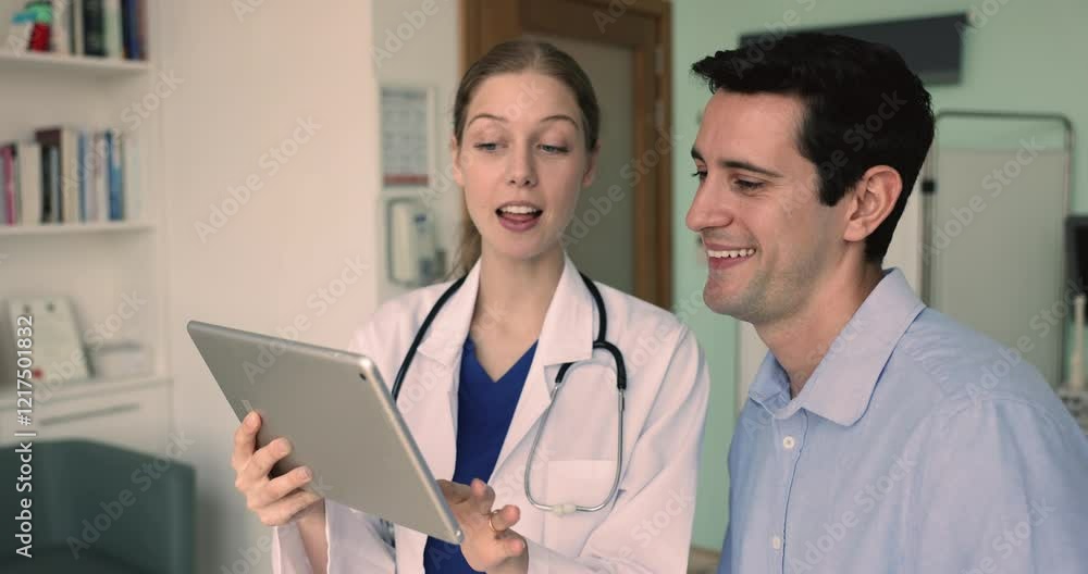 Happy young female doctor and male Hispanic patient watching online content on tablet. Medical practitioner showing and explaining checkup test results to patient man in examination room Happy young female doctor and male Hispanic patient watching online content on tablet. Medical practitioner showing and explaining checkup test results to patient man in examination room