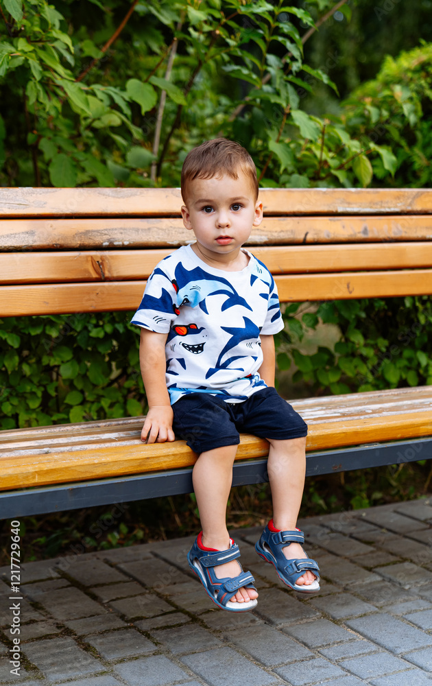 Child on a wooden bench outside. A young boy sits on a wooden bench in a green park, wearing a patterned shirt and shorts. Child on a wooden bench outside. A young boy sits on a wooden bench in a green park, wearing a patterned shirt and shorts.