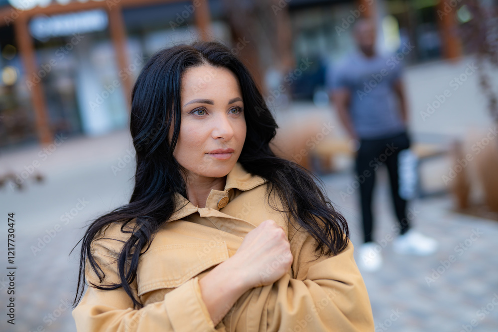 Fototapeta premium Woman in beige coat outdoors. A woman in a beige coat stands confidently, lost in thought, with a blurred figure behind her in the city.