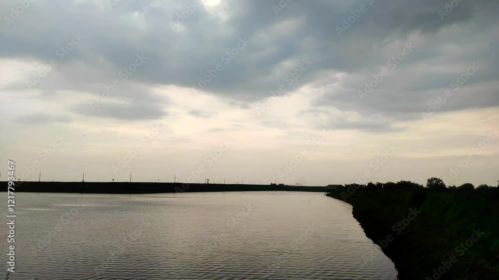 A serene dam with calm waters reflecting the cloudy sky, bordered by lush greenery and distant power poles, under an overcast atmosphere. A serene dam with calm waters reflecting the cloudy sky, bordered by lush greenery and distant power poles, under an overcast atmosphere.