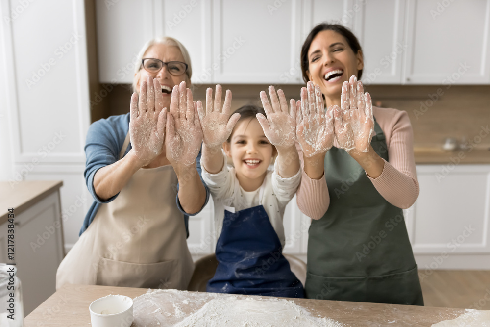 © fizkes - Having fun on kitchen. Cute family portrait laughing 3 female generations mature granny young mommy little tween child girl knead dough bake cake pastries together look at camera show palms in flour