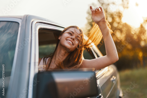 Happy young woman enjoying a road trip with her hair blowing in the wind, smiling from a car window during a sunny day in nature.