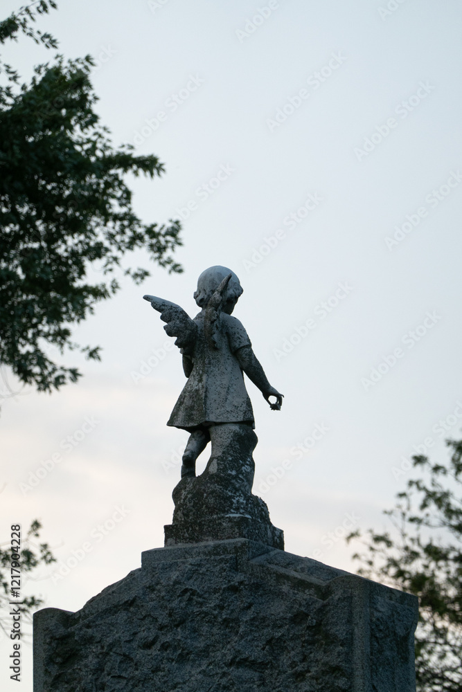 Fototapeta premium A weathered angel statue stands atop a stone pedestal, surrounded by trees. The figure, with delicate wings, captures a sense of timeless grace and quiet reflection against the backdrop of a clear sky