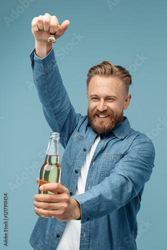 Cheerful bearded blonde hipster man open a bottle of beer over blue background in studio.