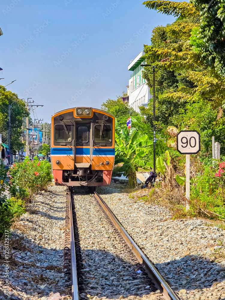 Naklejka premium A colorful train approaches a quiet station laden with lush greenery in Bangkok. The sun shines brightly, illuminating the railway tracks that run through a vibrant neighborhood.