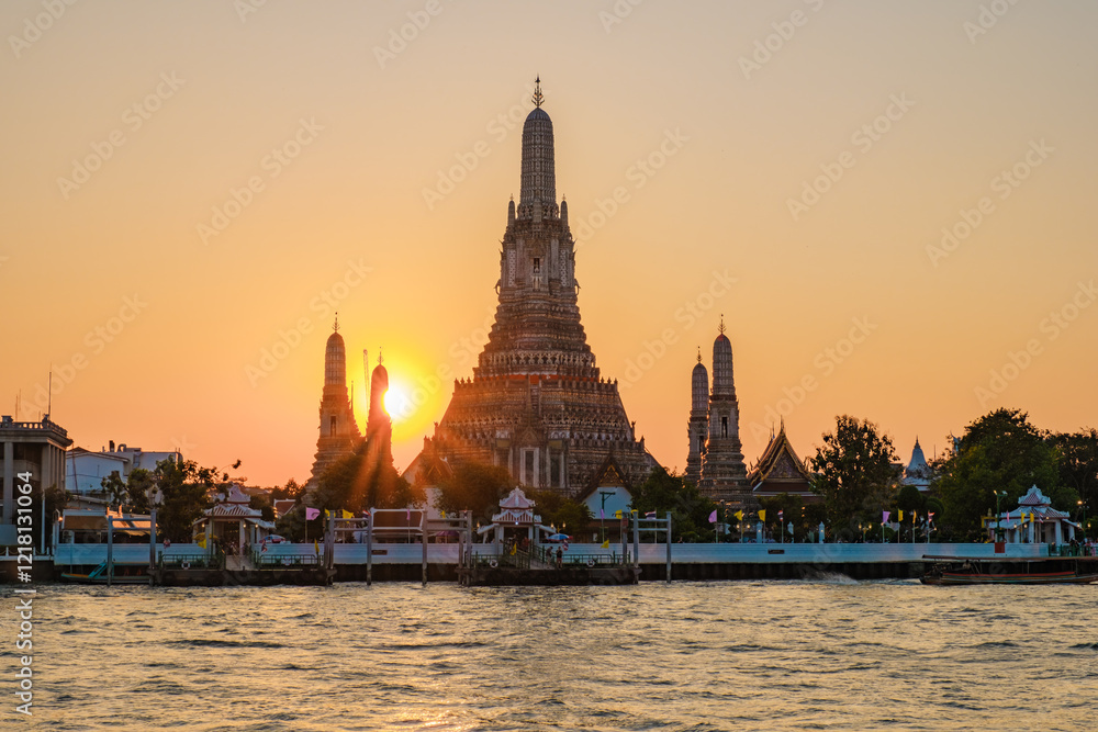 Fototapeta premium As the sun sets behind Wat Arun, its ornate spires glow with warm golden light, casting beautiful reflections on the tranquil waters of the Chao Phraya River. A serene moment in Bangkok.