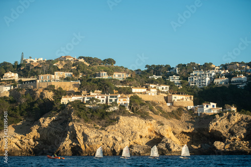 A peaceful scene of four small sailboats sailing in the clear waters of Mallorca, with rocky cliffs and charming houses dotting the backdrop