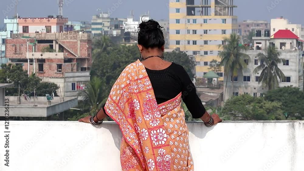 A Bengali woman in a beautiful floral cotton saree stands alone on a rooftop on a sunny day, looking out at the outdoor environment. Back view. A Bengali woman in a beautiful floral cotton saree stands alone on a rooftop on a sunny day, looking out at the outdoor environment. Back view.