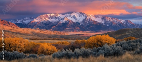 Majestic snow-capped mountains bathed in warm sunrise hues with vibrant orange and gold foliage in the foreground against a serene sky.