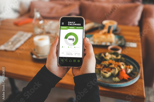Hands holding smartphone displaying daily calorie tracker app with nutritional information. Table with plates of food and a drink is blurred in background.