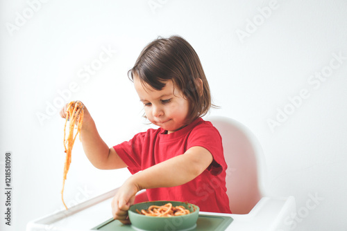 Little girl about 2 years old in a red shirt playing with spaghetti while eating from a green bowl in a highchair against a white background concept of childhood and fun meals photo