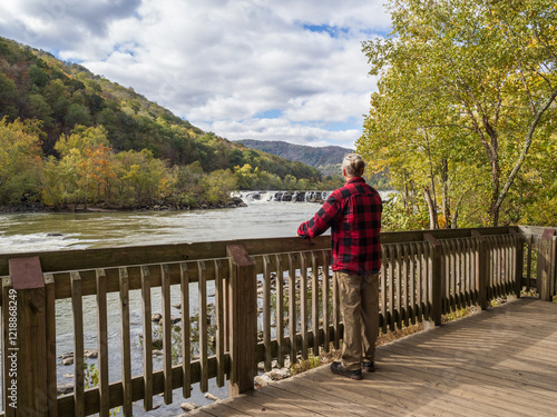 A man with a hair bun and red lumberjack jacket admires the vibrant fall colors at Sandstone Falls in New River Gorge National Park, West Virginia.