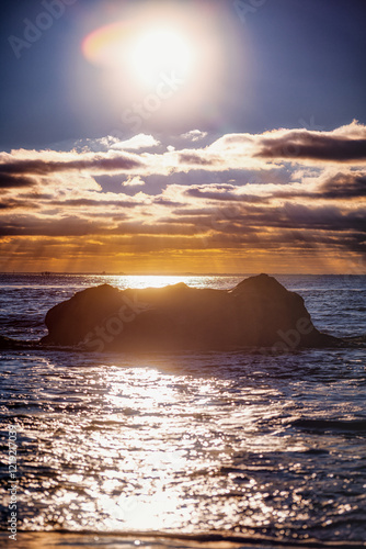 Un magnifique coucher de soleil illuminant la mer, avec un rocher en premier plan créant une silhouette contrastée sur l'eau scintillante.