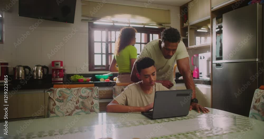Father assisting the young boy with his laptop at the dining table while the teenage daughter stands nearby drinking water in a cozy and busy family kitchen Father assisting the young boy with his laptop at the dining table while the teenage daughter stands nearby drinking water in a cozy and busy family kitchen