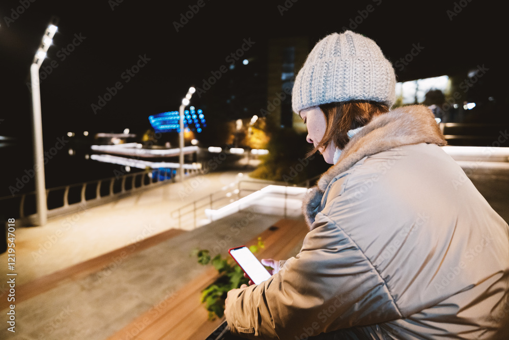 A woman in a light coat and a warm hat sits with a phone against the backdrop of an evening city A woman in a light coat and a warm hat sits with a phone against the backdrop of an evening city