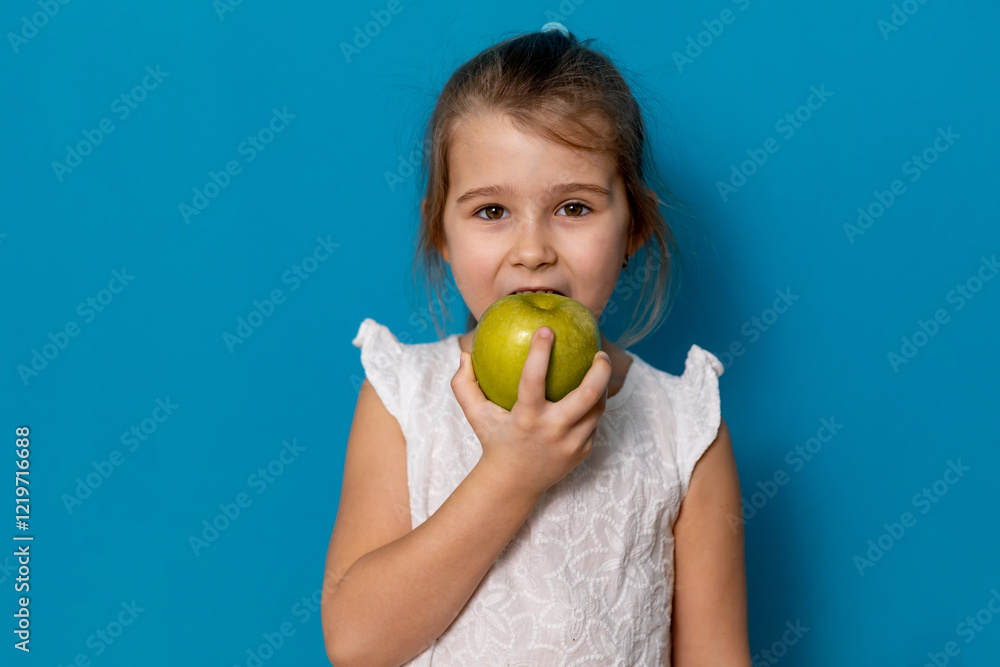 Cute child smiling bites apple. nutrition for caries prevention. portrait of little girl with fruit on blue background. kid eating healthy food, snack. teeth, dental health, smile. children dentistry Cute child smiling bites apple. nutrition for caries prevention. portrait of little girl with fruit on blue background. kid eating healthy food, snack. teeth, dental health, smile. children dentistry